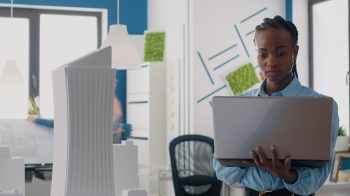Close up of engineer looking at laptop to analyze building model in architectural office. Woman builder using computer and maquette to design construction layout for development.